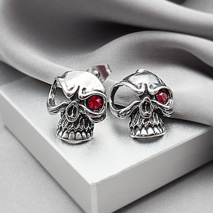 May include: A pair of silver-toned metal stud earrings shaped like skulls. Each skull features a single deep red circular gemstone set into one eye socket, with a polished metallic finish and detailed facial features including teeth and brow.