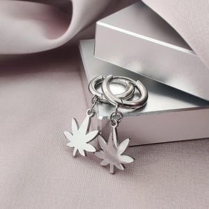 May include: A pair of silver-toned hoop earrings featuring dangling charms shaped like seven-pointed leaves. The earrings are constructed from a polished metallic material with a smooth, reflective surface, showcasing a simple and clean design.