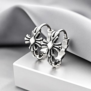May include: A pair of silver hoop earrings featuring a prominent, ornate cross design with oxidized detailing. The earrings have a circular hoop structure and a polished finish, showcasing intricate metalwork on the front-facing cross emblem.