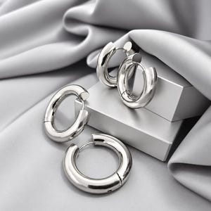 May include: A set of three silver-toned hoop earrings with a thick, rounded profile and a polished, reflective finish. The earrings feature a hinged closure mechanism and are arranged in a cluster, showcasing their smooth, circular design.
