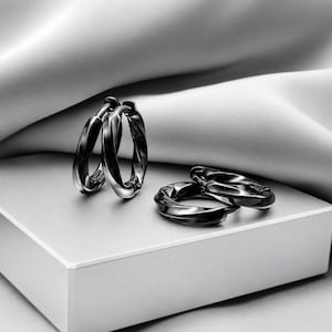 May include: A pair of polished black metal hoop earrings with a twisted, rope-like texture. One earring stands upright while the other lies flat, showcasing the reflective, dark metallic finish and the circular design of these jewelry pieces.