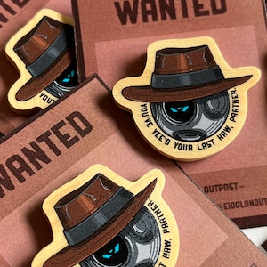 May include: A wooden pin featuring a cartoon robot wearing a brown fedora. The robot has a blue eye and the text "You've Yee'd Your Last Haw, Partner." is printed around the robot.