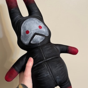 May include: A black plush bunny with red ears and red accents. The bunny has a white face with two red eyes and a black mouth.