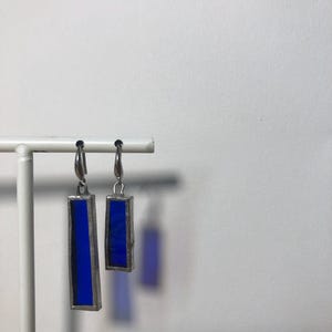 以下が含まれることがあります： 銀色の金属フレームに収められた深い青色の長方形のガラスピースが特徴のドロップイヤリングのペア。各イヤリングはシンプルな銀色のフックから吊り下げられており、このセットでは片方のイヤリングがもう片方よりも明らかに長くなっています。