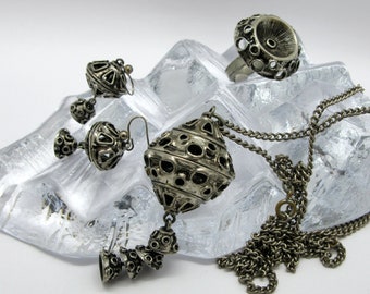 May include: A set of silver-toned jewelry featuring a pendant necklace, earrings, and a ring. The pendant is a large, ornate, round shape with a smaller, round shape hanging from it. The earrings are similar in design to the pendant, but smaller. The ring is a simple band with a large, round, ornate top.