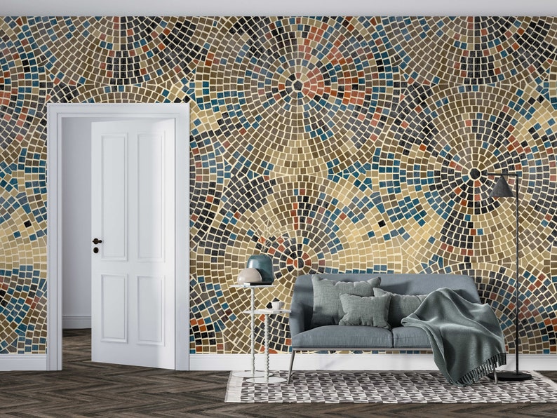 May include: A living room interior design featuring a wall covered in a mosaic pattern wallpaper with blue, brown, and beige tiles. A gray couch with pillows and a throw blanket is placed in front of the wall. A side table with a lamp and a floor lamp are also visible.