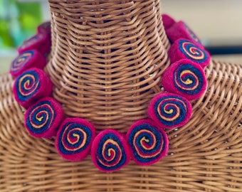 Könnte beinhalten: Eine Halskette aus Filz mit einer rosa Basis und einem mehrfarbigen Spiraldesign. Die Halskette wird auf einer Weidenpuppe präsentiert.