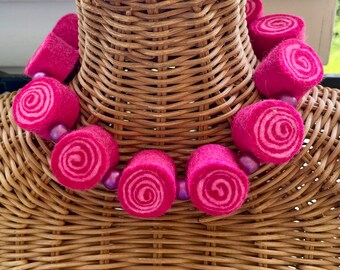 Könnte beinhalten: Eine rosafarbene Filzkette mit Spiralmuster. Die Kette besteht aus mehreren Filzrollen mit einer lila Perle zwischen jeder Rolle.