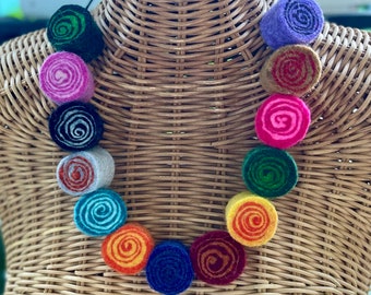 May include: A colorful felt necklace with a spiral design. The necklace features a variety of colors, including green, purple, pink, blue, orange, and yellow. The necklace is displayed on a wicker mannequin.