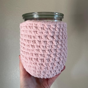 May include: A clear glass jar with a silver lid is encased in a soft, light pink crocheted cosy. The cosy has a textured, looped pattern. The jar is held in a person's hand.
