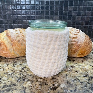 May include: White crocheted jar cosy with a glass jar inside. Two loaves of sourdough bread are on either side of the jar.