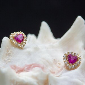 Op de afbeelding: Een paar hartvormige oorstekers. Elke oorbel heeft een levendige roze edelsteen in het midden, omringd door een halo van kleinere, heldere edelstenen. De oorbellen zijn gezet in goudkleurig metaal, wat een klassieke en elegante uitstraling creëert. De oorbellen worden op een wit oppervlak gepresenteerd.
