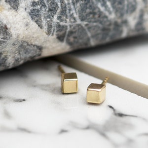 Könnte beinhalten: Ein Paar goldene Würfel-Ohrstecker. Die Ohrringe sind klein und schlicht und würden ein tolles Accessoire für jeden Tag sein.