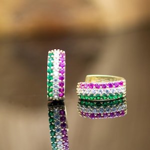 Puede incluir: Dos pendientes de aro dorados, cada uno incrustado con filas de piedras preciosas de colores. Un pendiente está de pie, mientras que el otro está acostado de lado. Las piedras preciosas están dispuestas en bandas verdes, blancas y moradas.
