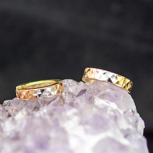 Puede incluir: Dos anillos de oro con un patrón geométrico se exhiben sobre un cristal púrpura. Los anillos tienen un diseño facetado, que refleja la luz. Uno es dorado y el otro es una mezcla de oro, plata y oro rosa. El fondo es oscuro.