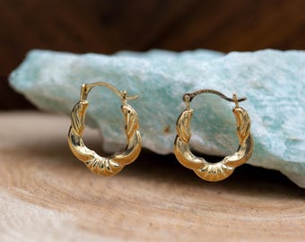 May include: A pair of gold-colored hoop earrings with a twisted rope design. The earrings are oval-shaped and have a textured surface. They are displayed on a wooden surface with a light blue stone in the background.