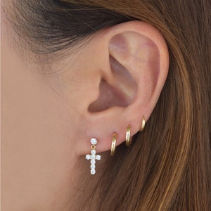Puede incluir: Aretes de aro de oro en varios tamaños, usados en el lóbulo superior de una oreja. Un pequeño arete de cruz colgante con piedras de circonita cúbica transparentes se usa en el lóbulo inferior.