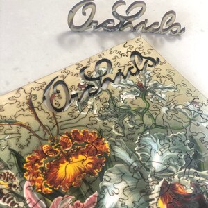 May include: A close-up of a jigsaw puzzle featuring a vintage illustration of orchids. The puzzle pieces are cut in a unique, intricate pattern. The word "Orchids" is spelled out in wooden letters above the puzzle.