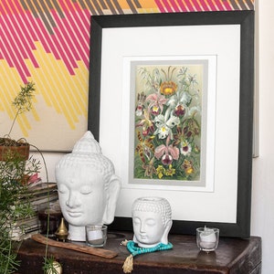 May include: A framed botanical print of white and pink orchids with green leaves against a light beige background. The print is displayed on a wooden chest with two white ceramic Buddha heads, a small gold incense burner, and teal blue beads.