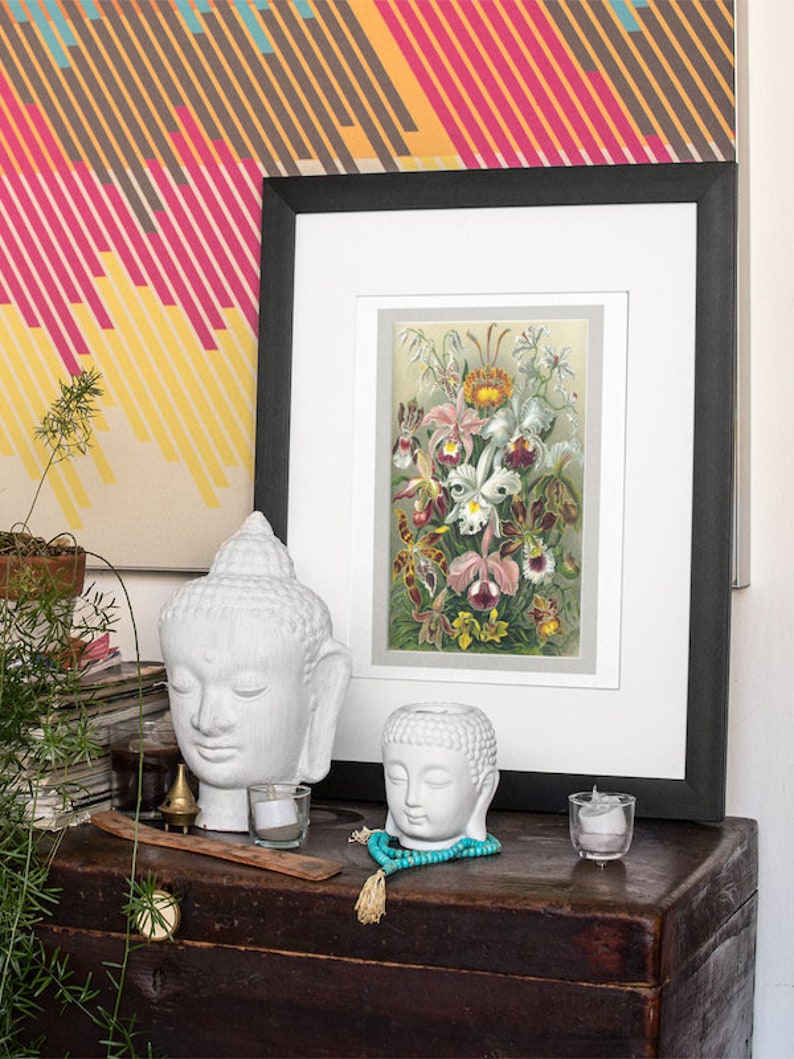 May include: A framed botanical print of white and pink orchids with green leaves against a light beige background. The print is displayed on a wooden chest with two white ceramic Buddha heads, a small gold incense burner, and teal blue beads.