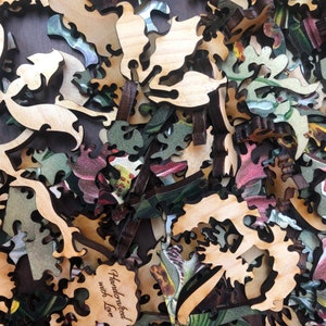 May include: A close-up of a pile of wooden jigsaw puzzle pieces with a floral pattern. The pieces are cut in intricate shapes and have a natural wood finish. One piece has the text "Handcrafted with Love" engraved on it.