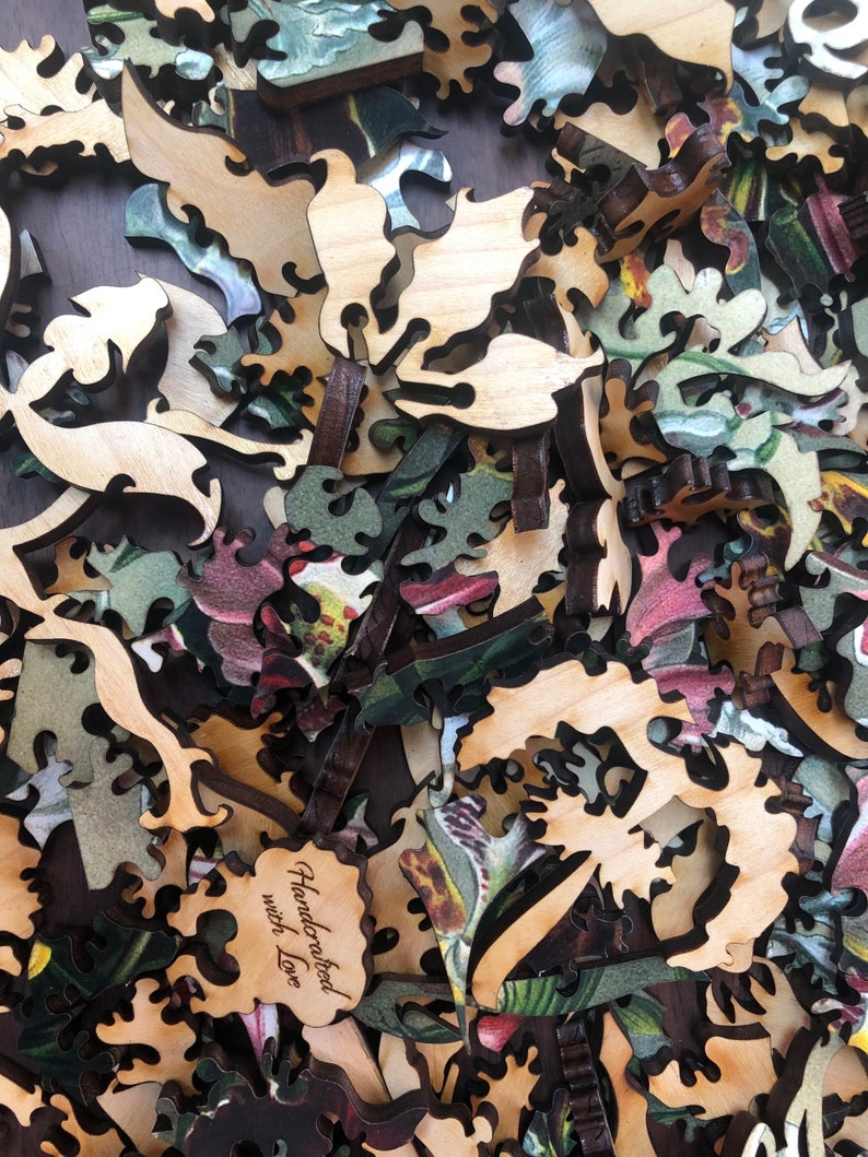 May include: A close-up of a pile of wooden jigsaw puzzle pieces with a floral pattern. The pieces are cut in intricate shapes and have a natural wood finish. One piece has the text "Handcrafted with Love" engraved on it.