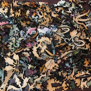 May include: A close-up of a wooden jigsaw puzzle with intricate pieces in various shapes and sizes. The pieces are scattered on a dark brown surface. The puzzle features a floral design with a variety of colors, including green, blue, pink, and yellow. The text "Handcrafted with love" is visible on one of the pieces.