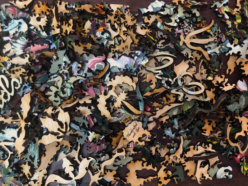 May include: A close-up of a wooden jigsaw puzzle with intricate pieces in various shapes and sizes. The pieces are scattered on a dark brown surface. The puzzle features a floral design with a variety of colors, including green, blue, pink, and yellow. The text "Handcrafted with love" is visible on one of the pieces.