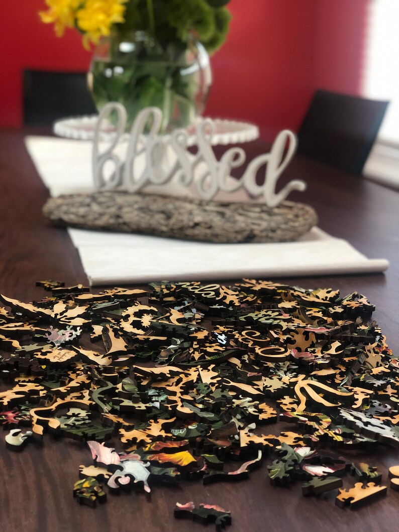 May include: A wooden jigsaw puzzle with a floral design, scattered on a dark wooden table. The puzzle pieces are in various shades of brown, green, and yellow. The word "blessed" is visible in the background.