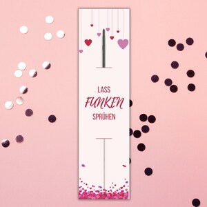 Puede incluir: Una tarjeta blanca con el texto "Lass Funken Sprühen" y corazones rosas colgando de la parte superior. La tarjeta está rodeada de confeti plateado y negro sobre un fondo rosa.