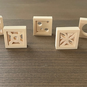 Puede incluir: Cinco pequeños bloques cuadrados de madera de color claro con varios diseños recortados. Los diseños incluyen círculos, patrones geométricos y motivos florales. Los bloques están dispuestos sobre una superficie de madera oscura.