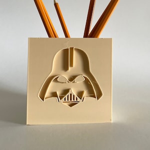 May include: A beige pencil holder shaped like Darth Vader's head. The holder has a 3D printed design and is holding several yellow pencils.