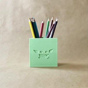 May include: A light green pencil holder with a 3D printed design of a character from the Star Wars franchise. The character is a small, green, humanoid creature with large ears and eyes. The holder is filled with colored pencils.