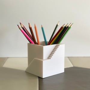 May include: A white, geometric pencil holder with a staircase design. The holder is filled with a variety of colored pencils, including pink, brown, orange, and green. The pencils are arranged in a neat and organized manner.