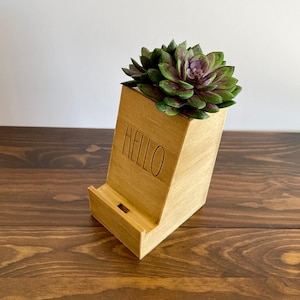 Puede incluir: Soporte de teléfono de madera con una planta suculenta en una maceta. El soporte tiene la palabra "HELLO" grabada en la parte delantera.