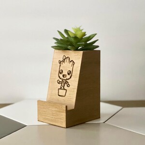 Peut inclure: Un support de téléphone en bois et un pot à succulentes avec une image gravée au laser d'un personnage de dessin animé Groot. Le pot est de couleur brun clair et le support de téléphone est de couleur brun foncé.