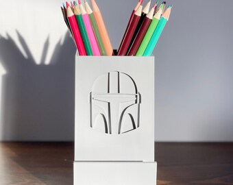 May include: White pencil holder with a Mandalorian helmet design. The holder is filled with colored pencils.
