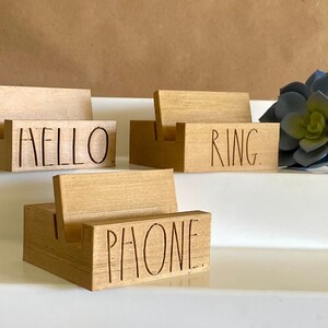 May include: Three wooden phone stands with the words "HELLO", "RING", and "PHONE" engraved on them. The stands are arranged in a staggered formation on a white surface. A blue succulent plant is in the background.