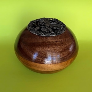 Peut inclure: Un urne en bois faite à la main avec une finition marron foncé et une base en bois plus clair. L'urne est surmontée d'un couvercle décoratif en métal avec un motif d'oiseau gravé. L'urne a une forme lisse et arrondie.
