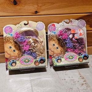 Könnte beinhalten: Zwei Packungen mit Cabbage Patch Kids Haarschmuck. Jede Packung zeigt einen Puppenkopf mit verschiedenen Frisuren und Accessoires. Der Text auf den Packungen lautet "Cabbage Patch Kids" und "Accessory Accessories".