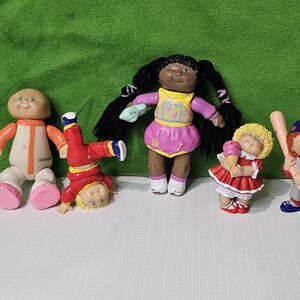 Peut inclure: Cinq poupées Cabbage Patch Kids, chacune avec une tenue et une coiffure différentes. Les poupées portent toutes des tenues différentes, dont un uniforme de baseball rouge et blanc, une robe rose avec un cornet de glace jaune, une combinaison rouge et blanche, une robe rose avec une fleur jaune et verte, et une combinaison rouge et blanche.