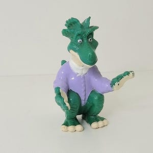 Może przedstawiać: Zielona i fioletowa plastikowa figurka dinozaura. Dinozaur ma na sobie fioletową koszulę i ma zielone ciało z białym brzuchem. Dinozaur siedzi na swoich zadach z wyciągniętymi ramionami.