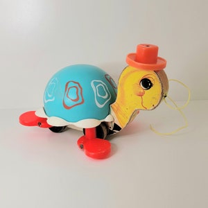Peut inclure: Une tortue jouet vintage avec une carapace bleue et des pieds rouges. La tortue a une tête jaune avec un petit chapeau orange et une ficelle attachée. La carapace a un motif blanc et orange.