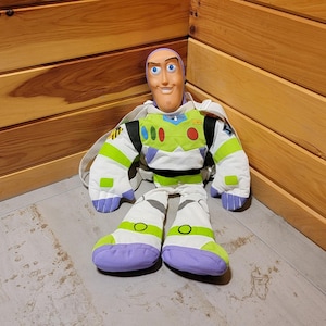May include: A plush Buzz Lightyear backpack with a green and white spacesuit, purple boots, and a smiling face. The backpack is sitting on a wooden floor.