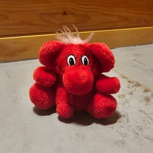 Puede incluir: Un elefante de peluche rojo con pelo blanco en la cabeza y ojos negros. El juguete tiene una expresión sonriente.