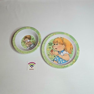 Peut inclure: Un ensemble de vaisselle pour enfants en plastique vert et blanc avec un motif de dessin animé représentant une fille. La fille porte une chemise blanche et une jupe bleue avec une coccinelle rouge dans la main. L'assiette et le bol ont une bordure verte avec des fleurs blanches et des feuilles vertes.