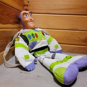 May include: A plush Buzz Lightyear toy from the Toy Story franchise. The toy is wearing a green and white spacesuit with a purple chest piece and a large, cartoonish face. The toy is sitting on a light gray surface.