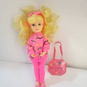 May include: A small doll with blonde curly hair wearing a pink sweater with a geometric pattern, pink trousers, and pink shoes. The doll is holding a pink handbag with a geometric pattern.