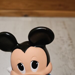 May include: A close-up of a Mickey Mouse figurine. The figurine has black ears, a tan face, and a black nose. It is wearing a blue shirt with yellow and red accents. The eyes are black and the mouth is a dark color.