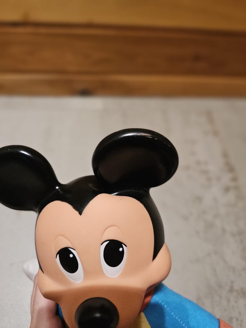 May include: A close-up of a Mickey Mouse figurine. The figurine has black ears, a tan face, and a black nose. It is wearing a blue shirt with yellow and red accents. The eyes are black and the mouth is a dark color.